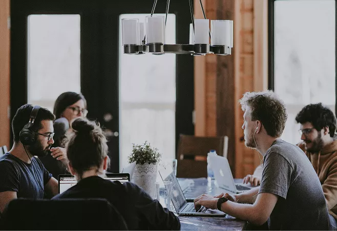 Five team members work on laptops around a wooden table in a warmly lit collaborative workspace.