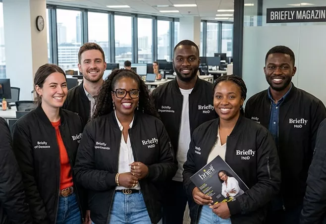 Six Briefly Magazine team members in branded black jackets smile together in their open-plan office.